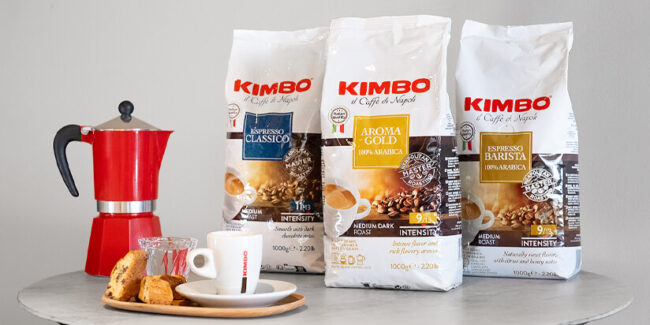 Kimbo Espresso Kaffeeverpackungen auf einem runden Tisch. Daneben ein roter Espressokocher und weiße Espressotasse.