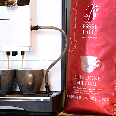 Essse Selezione Speciale aus dem Kaffeevollautomat Espresso fließt aus Kaffeevollautomat in zwei Espressotassen. Daneben rote Verpackung Essse Selezione Speciale