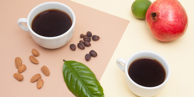 Zwei Kaffeetassen mit schwarzem Kaffee, Mandeln, Kaffeebohnen, Granatapfel, Limette und ein Blatt liegen auf einem Hintergrund