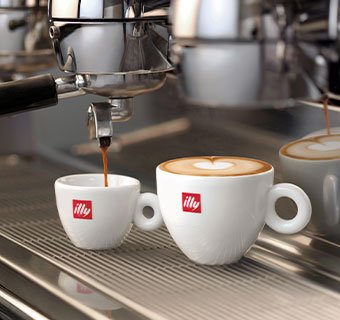 Espresso läuft aus der Siebträgermaschine in eine weiße illy Tasse, daneben steht ein Cappuccino  mit Latte Art