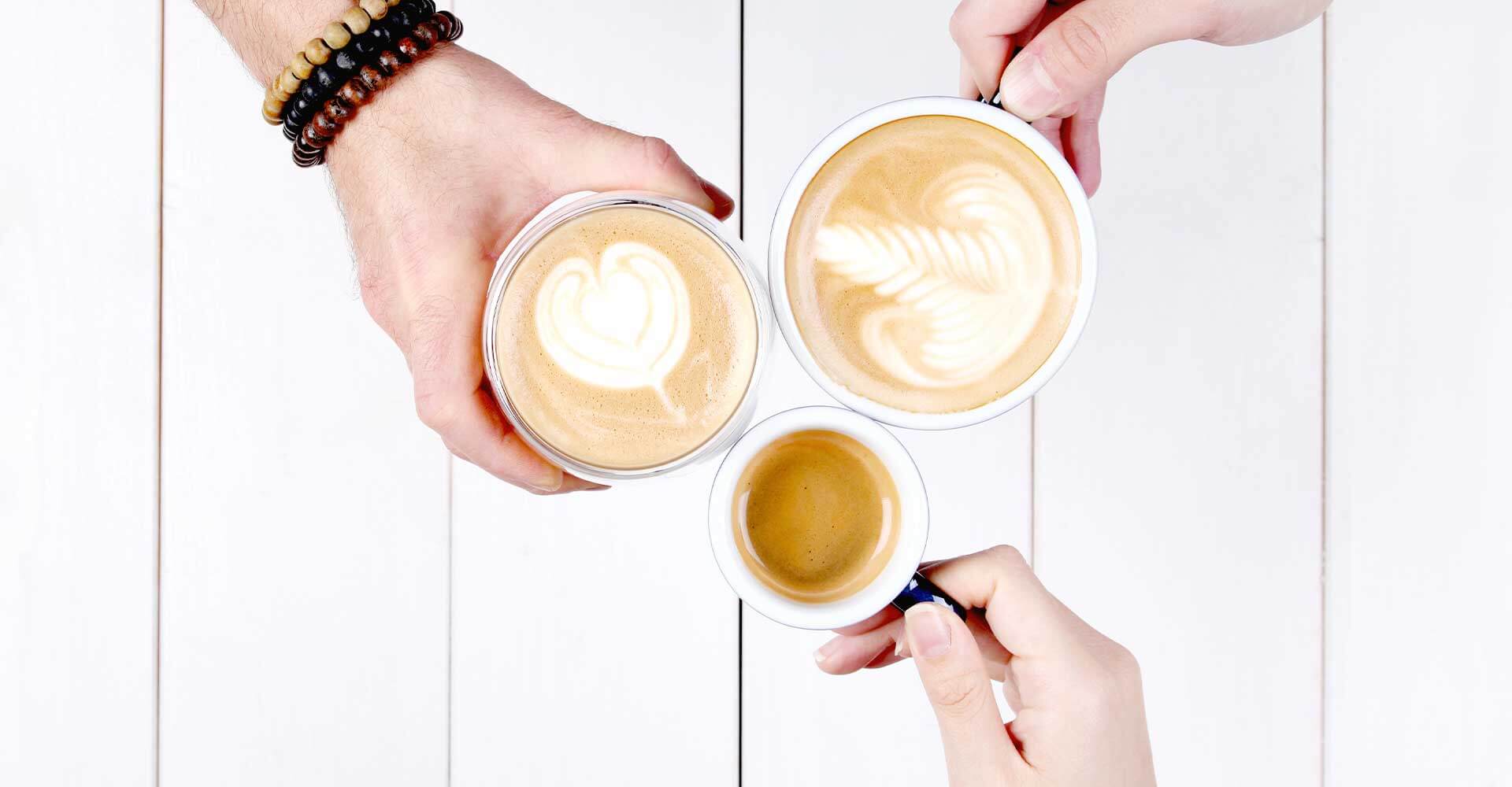 Drei Tassen stoßen miteinander an, eine Espressotasse, ein Cappuccino mit Latte Art Herz sowie ein großer Milchkaffee mit Latte Art
