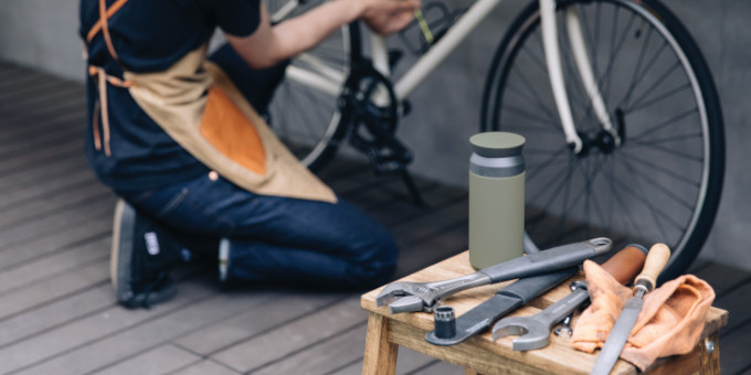 Werkzeug und grüner Kaffeebecher stehen auf Holzhocker. Im Hintergrund schraubt Person an einem Fahrrad